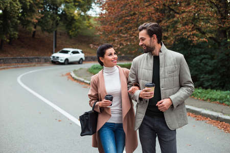 Perfect autumn walk. Young beautiful couple bonding to each other while walking in the park in autumn with coffee cupsの写真素材