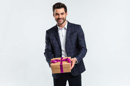 Your birthday gift. Handsome young man holding a present box and looking at camera with smile while standing against white backgroundの写真素材