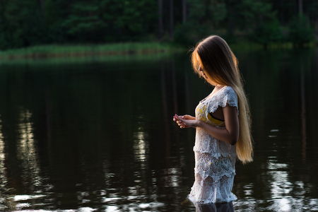Beautiful young girl in white dress standing in water on sunsetの写真素材