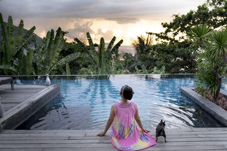 Intercontinental Peninsula Resort Da Nang, Vietnam - July 8, 2018: a woman and her pet dog Chihuahua was  sitting relax by the pool at sunset in Intercontinental Peninsula luxury resort Danang, Vietnamのeditorial素材