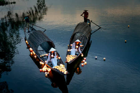 Hue City, Vietnam - May 8, 2020: Two Vietnamese girls with wihte tradition Ao Dai are lighting candle in lantern to pray in the river. Ao dai is famous traditional costume for the women in Vietnam.のeditorial素材
