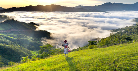 A young woman with a backpack is walking on a green hill among the clouds.