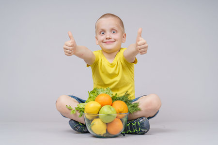 Smiling boy standing with fruits and vegetables in a plate, isolated on grayの写真素材