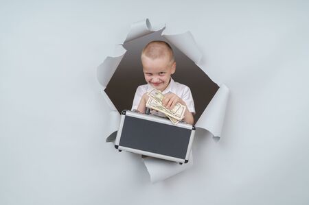 Cool boy is holding a suitcase full of money and emerging through torn paper hole in studio, has excited cheerful expression, looks through breakthrough of gray background.の写真素材