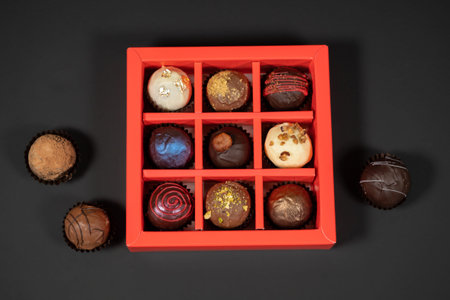 Various handmade chocolate truffle candies in a red box on a dark background. Directly above top...
