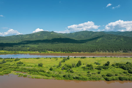 Flying above a glacial river in Siberia, Russia with green mountains and railway track in the...