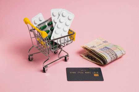 Pills in shopping cart, money and bank card on pink background. Concept of buying pills online
