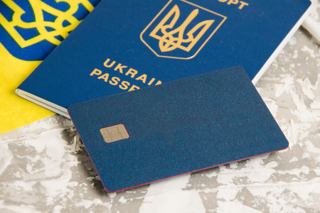 Bank card with chip close-up, Ukrainian passport and flag on gray concrete background.