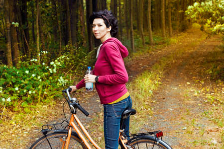 Young beautiful girl on a bicycle with a bottle of water in hand in the woods. Walking on a bicycle in the forest. A slim woman with a sports body shape on a bicycle.の写真素材