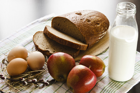 Country still life with food farming. Milk, bread, eggs, red apples and a sprig of willow on a towel on a black background. Easter still life.の写真素材