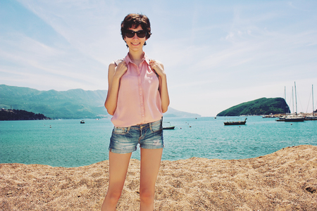 A smiling young girl tourist with a backpack and sunglasses standing by the sea on a background of mountains on a sunny day in vintage toning. Tourist on the beach. Travel photo of pretty woman.の写真素材