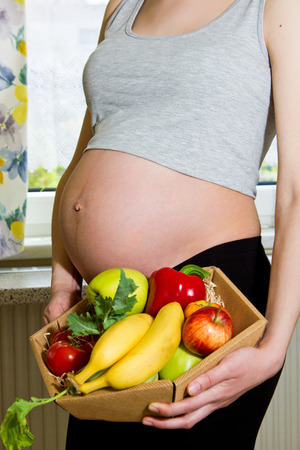 Young beautiful pregnant woman holding a basket of fruits and vegetables. A pregnant woman is eating in the kitchen.の写真素材