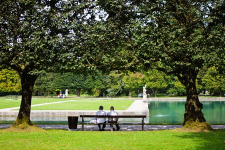 Elderly couple sits on a bench in the park of the Hellbrunn palace. The beautiful park with a pond and trees in which people walkの写真素材