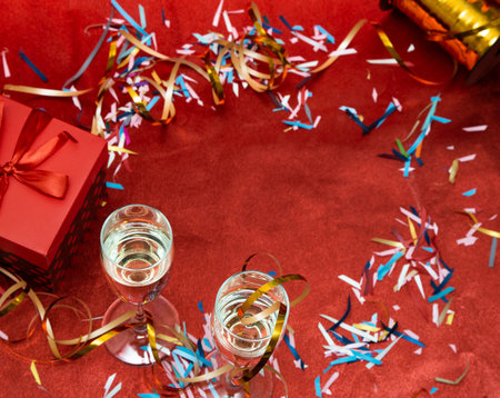 Two glasses with champagne and a gift box stand on a red background with confetti and serpentine. Greeting card for New Year and Christmas. Romantic postcard.の写真素材