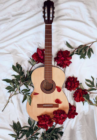 An acoustic wooden guitar lies on a white sheet surrounded by red peonies. Cover for music album, music concept, guitar playing, romantic serenadeの写真素材