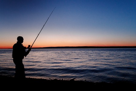 Silhouette of a fisherman with a fishing rod at a beautiful sunset. Against mountainsの写真素材