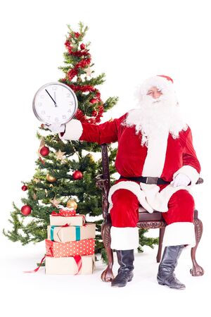 Portrait of Santa Claus sitting in chair next to Christmas tree holding clock, heap of giftboxes near himの写真素材