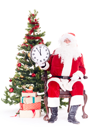 Portrait of Santa Claus sitting in chair next to Christmas tree holding clock, heap of giftboxes near himの写真素材