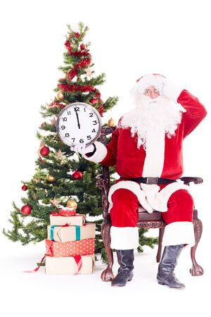 Portrait of Santa Claus sitting in chair next to Christmas tree holding clock, heap of giftboxes near himの写真素材