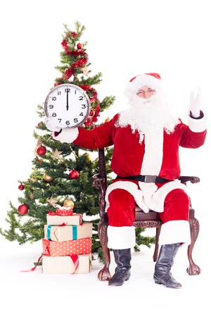 Portrait of Santa Claus sitting in chair next to Christmas tree holding clock, heap of giftboxes near himの写真素材