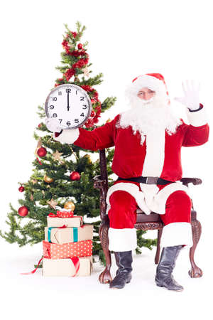 Portrait of Santa Claus sitting in chair next to Christmas tree holding clock, heap of giftboxes near himの写真素材