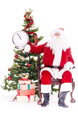 Portrait of Santa Claus sitting in chair next to Christmas tree holding clock, heap of giftboxes near himの写真素材