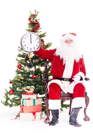 Portrait of Santa Claus sitting in chair next to Christmas tree holding clock, heap of giftboxes near himの写真素材
