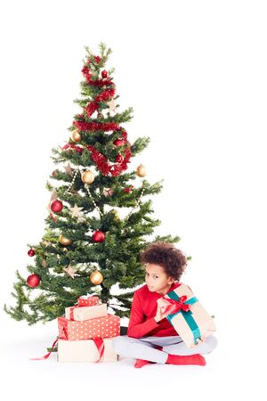 Portrait of cute mixed race little boy sitting on floor near Christmas tree with gift box in his handsの写真素材