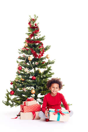 Portrait of cute mixed race little boy sitting on floor near Christmas tree with gift box in his handsの写真素材