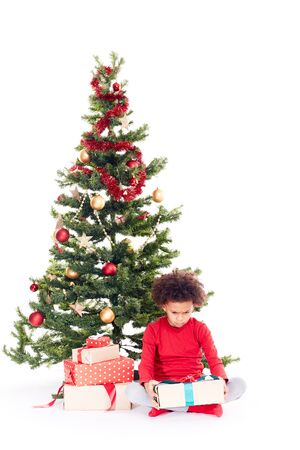 Portrait of cute mixed race little boy sitting on floor near Christmas tree with gift box in his handsの写真素材
