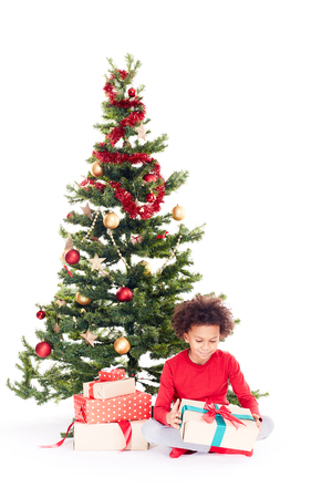 Portrait of cute mixed race little boy sitting on floor near Christmas tree with gift box in his handsの写真素材