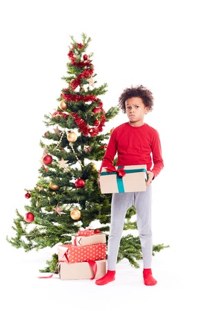 Portrait of cute mixed race little boy standing near Christmas tree with gift box in his handsの写真素材