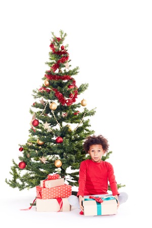Portrait of cute mixed race little boy sitting on floor near Christmas tree with gift box in his handsの写真素材