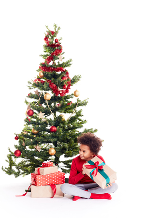 Portrait of cute mixed race little boy sitting on floor near Christmas tree with gift box in his handsの写真素材