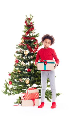 Portrait of cute mixed race little boy standing near Christmas tree with gift box in his handsの写真素材