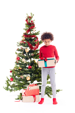 Portrait of cute mixed race little boy standing near Christmas tree with gift box in his handsの写真素材