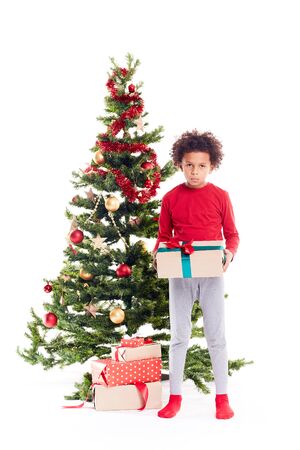 Portrait of cute mixed race little boy standing near Christmas tree with gift box in his handsの写真素材