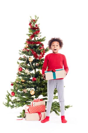 Portrait of cute mixed race little boy standing near Christmas tree with gift box in his handsの写真素材