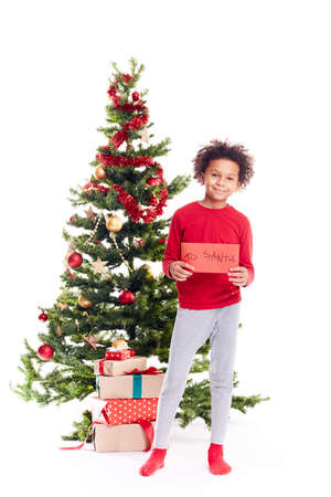 Portrait of cute mixed race little boy standing near Christmas tree holding letter to Santaの写真素材