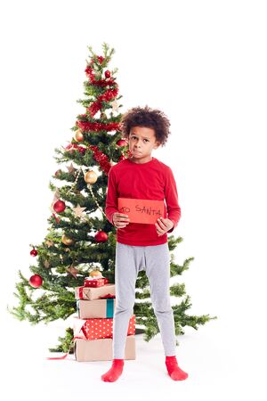 Portrait of cute mixed race little boy standing near Christmas tree holding letter to Santaの写真素材