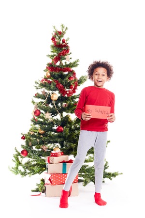 Portrait of cute mixed race little boy standing near Christmas tree holding letter to Santaの写真素材