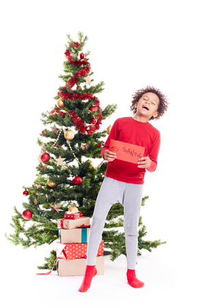 Portrait of cute mixed race little boy standing near Christmas tree holding letter to Santaの写真素材