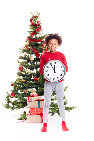 Portrait of cute mixed race little boy standing near Christmas tree holding clock showing five minutes to twelveの写真素材