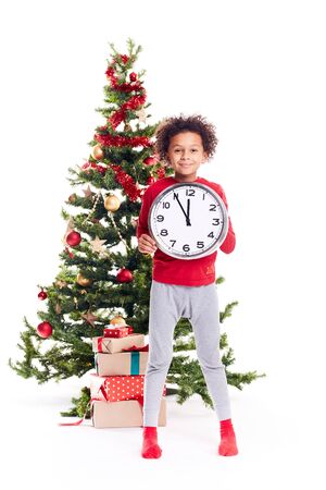 Portrait of cute mixed race little boy standing near Christmas tree holding clock showing five minutes to twelveの写真素材