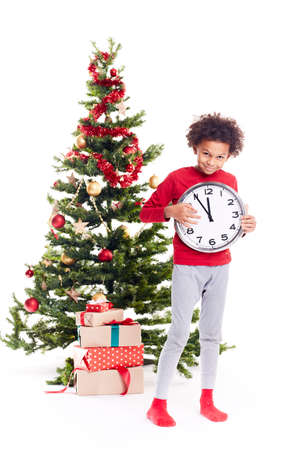 Portrait of cute mixed race little boy standing near Christmas tree holding clock showing five minutes to twelveの写真素材