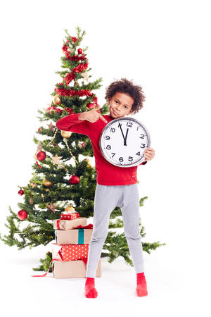 Portrait of cute mixed race little boy standing near Christmas tree holding clock showing five minutes to twelveの写真素材
