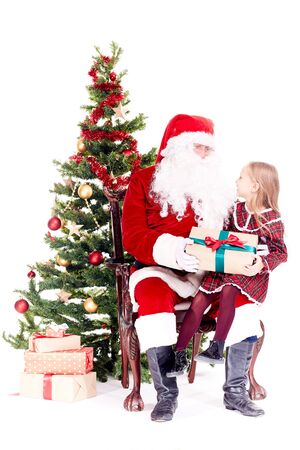 Cute little girl receiving gift from Santa Claus sitting on his lap near Christmas treeの写真素材