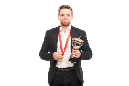 Portrait of confident young businessman wearing medal and holding trophy on white backgroundの写真素材