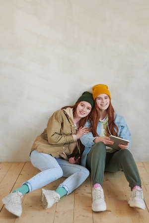 Vertical high angle studio portrait of joyful young female twins wearing stylish casual outfits sitting together on flloor smiling at cameraの写真素材