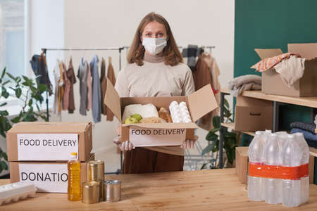 Unrecognizable woman wearing mask and latex gloves holding cardboard box with food supplies for donationの写真素材